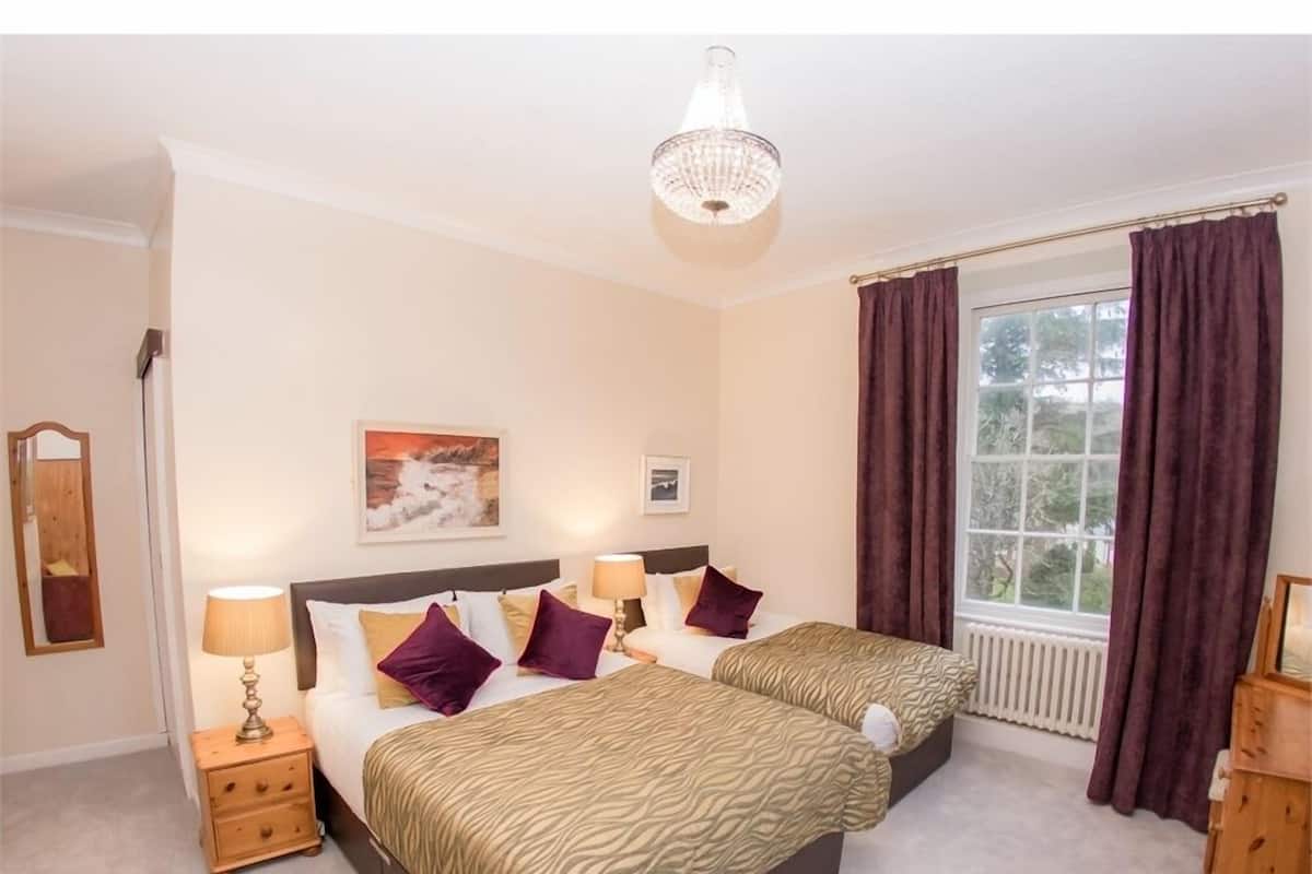 A hotel room with two beds, a nightstand with a lamp, a painting on the wall, and a window with curtains.