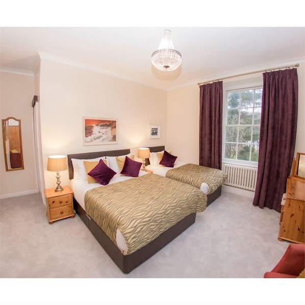 A hotel room with two beds, a nightstand with a lamp, a painting on the wall, and a window with curtains. - Royal Goat Hotel (Caernarfon)