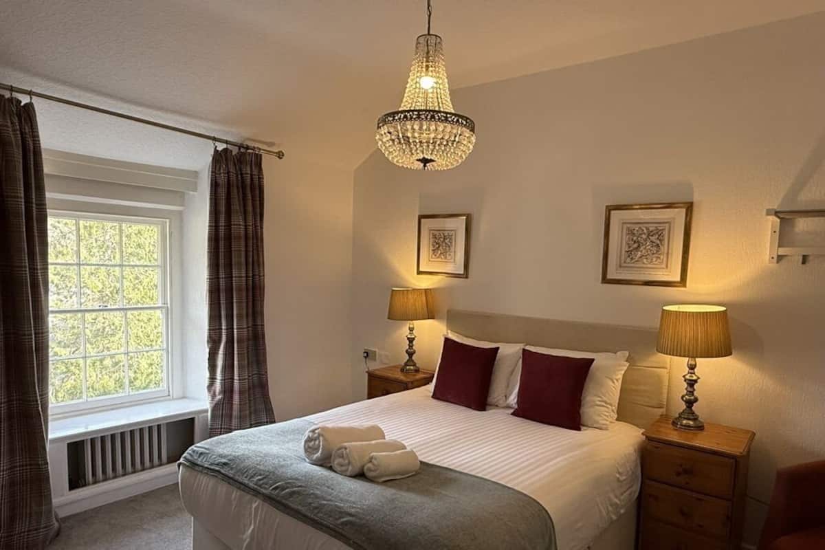A neatly made bed with a grey blanket and two towels, two bedside lamps, a chandelier, and two framed pictures on the wall.