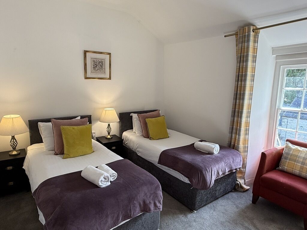 A hotel room with two beds, each with a pillow and a blanket, a nightstand with a lamp, a red sofa, and a window with curtains.
