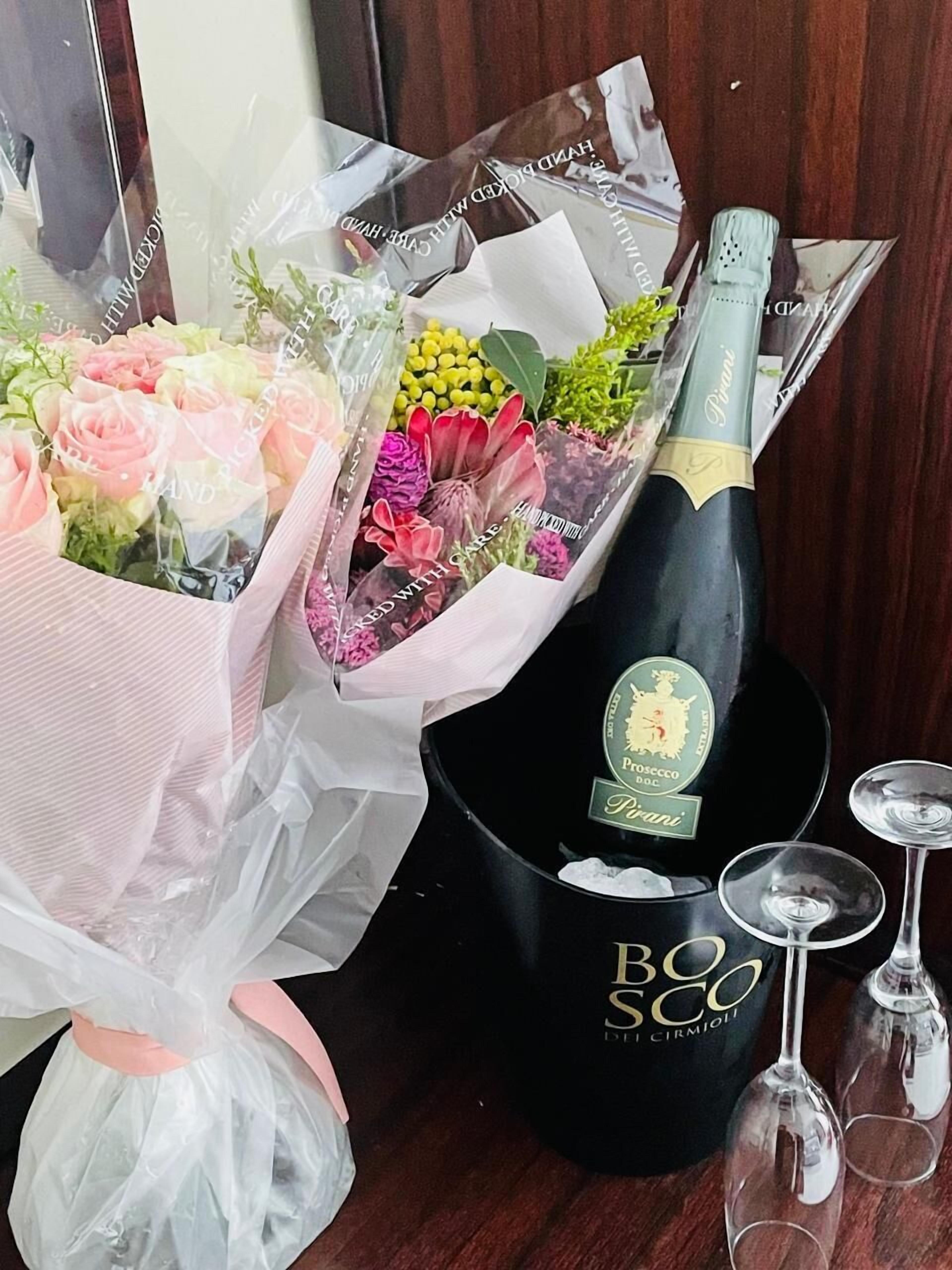 Une bouteille de vin BOSS DOC, un bouquet de fleurs et deux verres à vin posés sur une surface en bois.