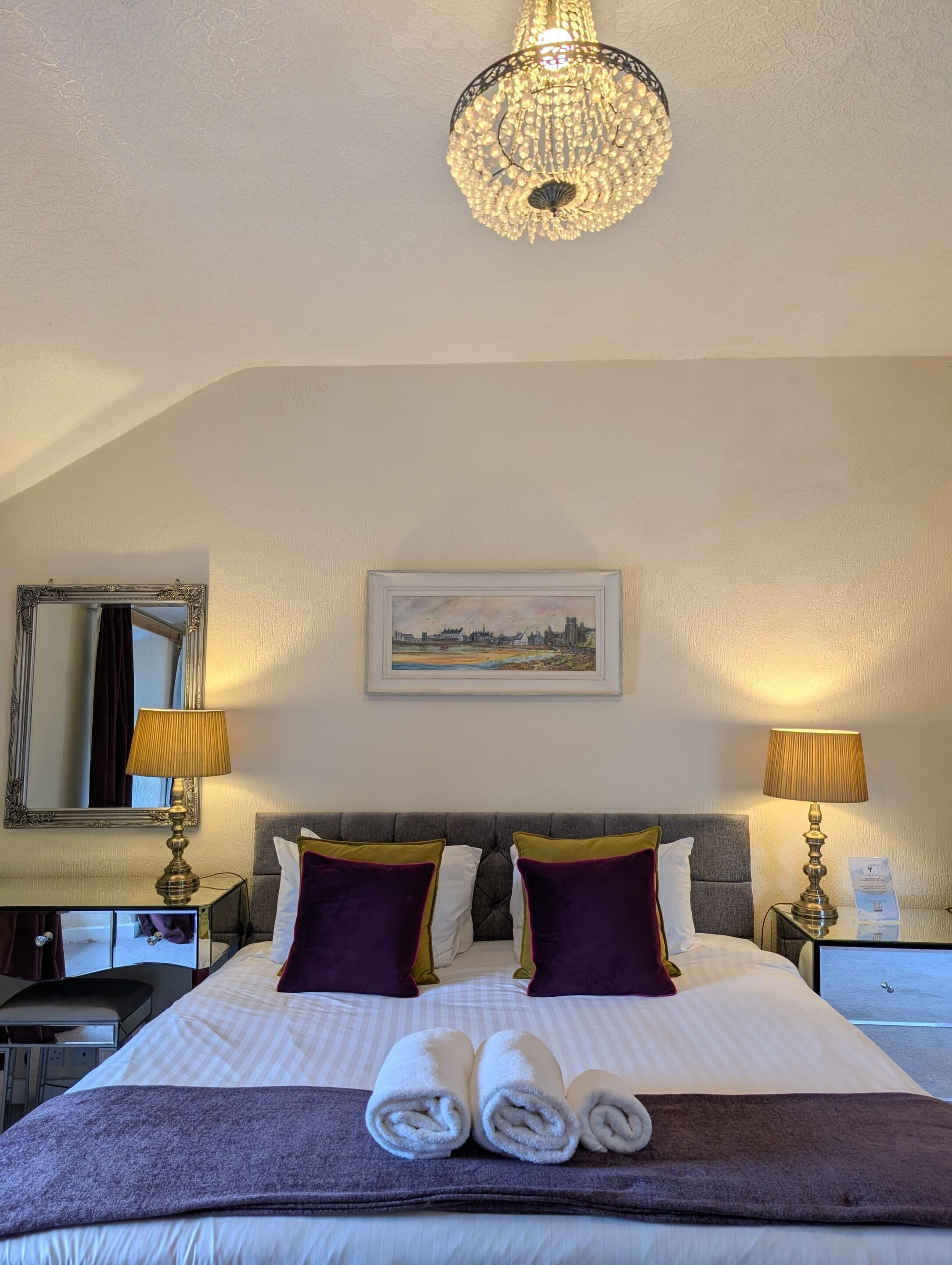 Un cuarto de hotel con una cama grande, dos lámparas de noche, un candelabro, un cuadro enmarcado de un paisaje urbano y un espejo en la pared.