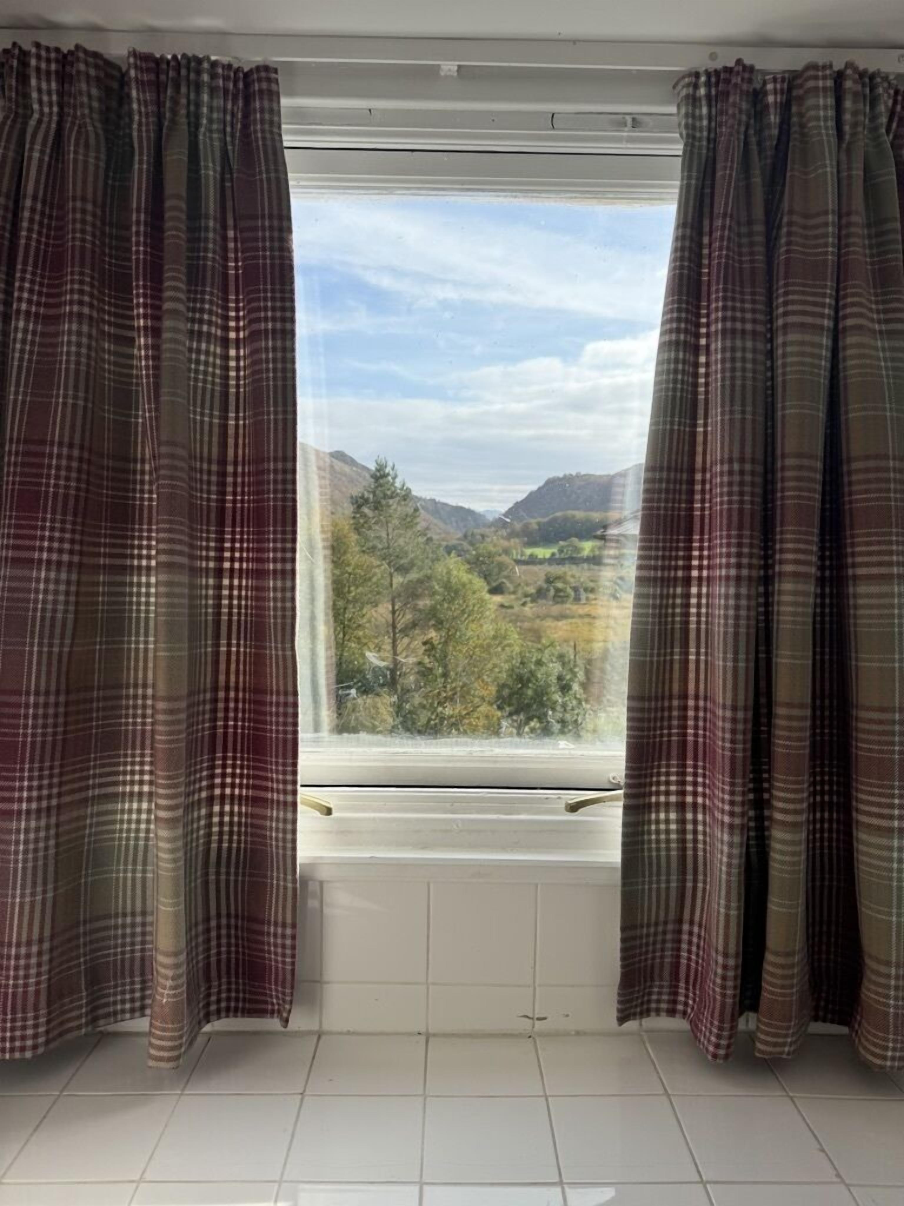 Una habitación con ventana y cortinas, piso de baldosas y vista a un paisaje montañoso.