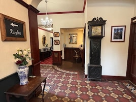 Un área de recepción con una alfombra de colores beige y rojo, un mostrador de recepción, un florero con flores, un reloj y cuadros en la pared.