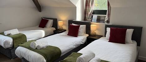 A hotel room with three single beds, each with a green throw blanket and white bedding. The room has a sloped ceiling with exposed beams and a window with curtains.