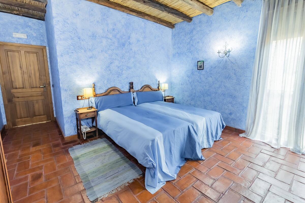A bedroom with a bed, wooden headboard, two bedside tables with lamps, a door, and a window with curtains.