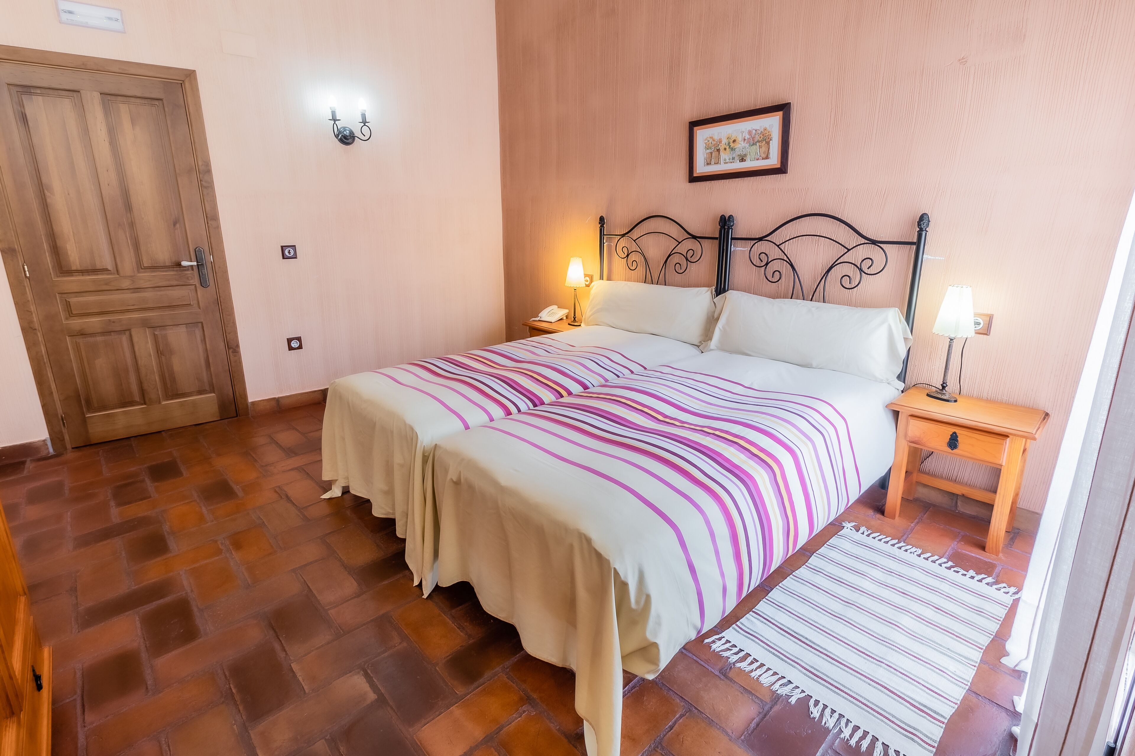 Un dormitorio con una cama, dos mesitas de noche, una puerta de madera y una lámpara fijada a la pared.