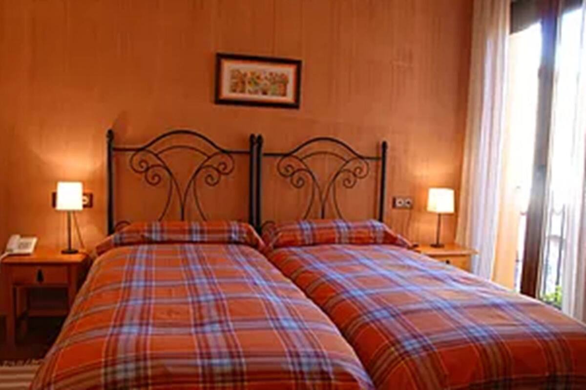 A double bed with a plaid bedspread, two bedside tables with lamps, a framed picture on the wall, and a window with curtains.