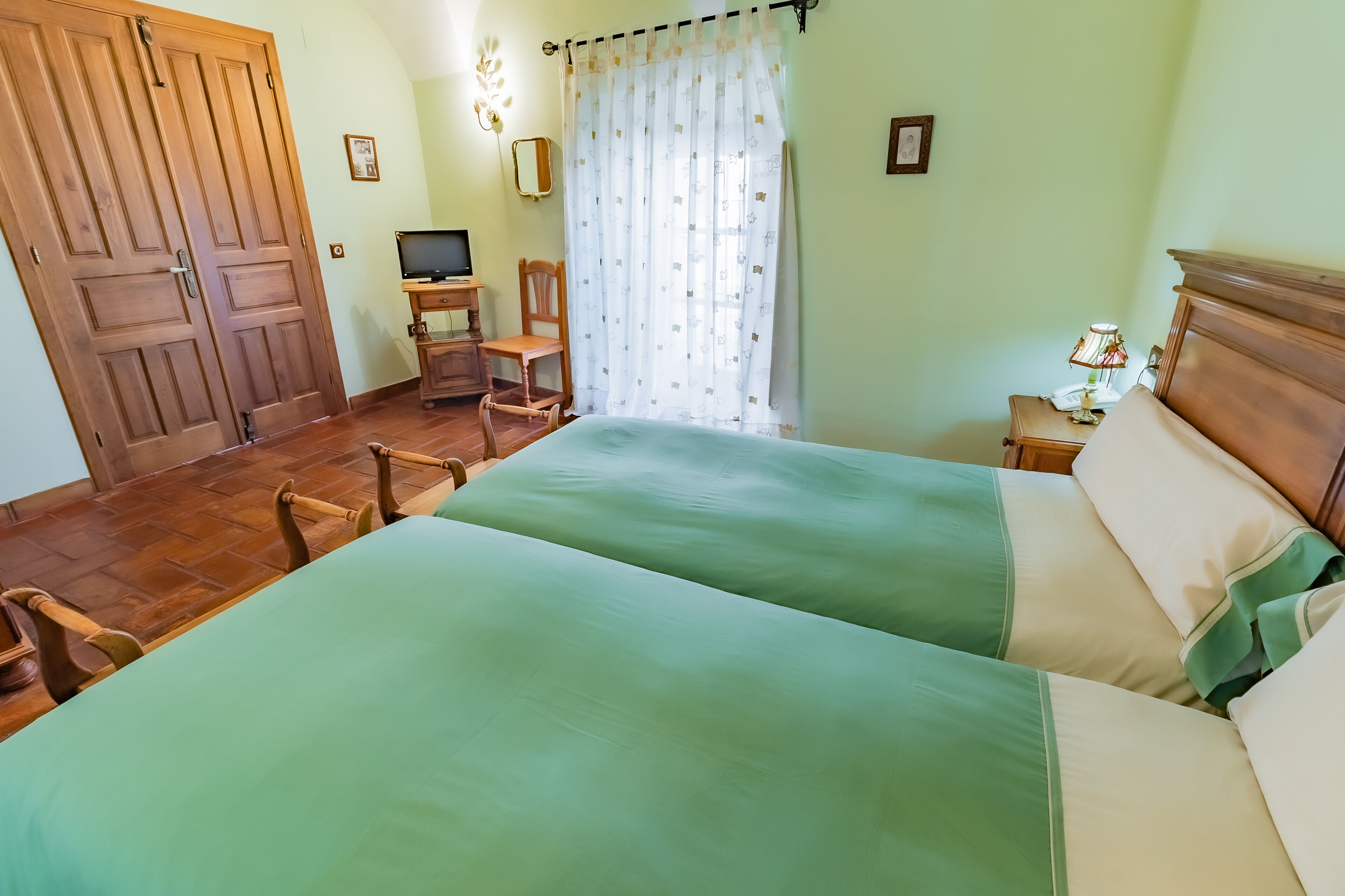 Una habitación con dos camas, un escritorio con computadora, una silla, una mesita de noche con lámpara y un ventanal con cortinas.