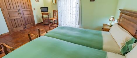 Una habitación con dos camas, un escritorio con computadora, una silla, una mesita de noche con lámpara y un ventanal con cortinas.