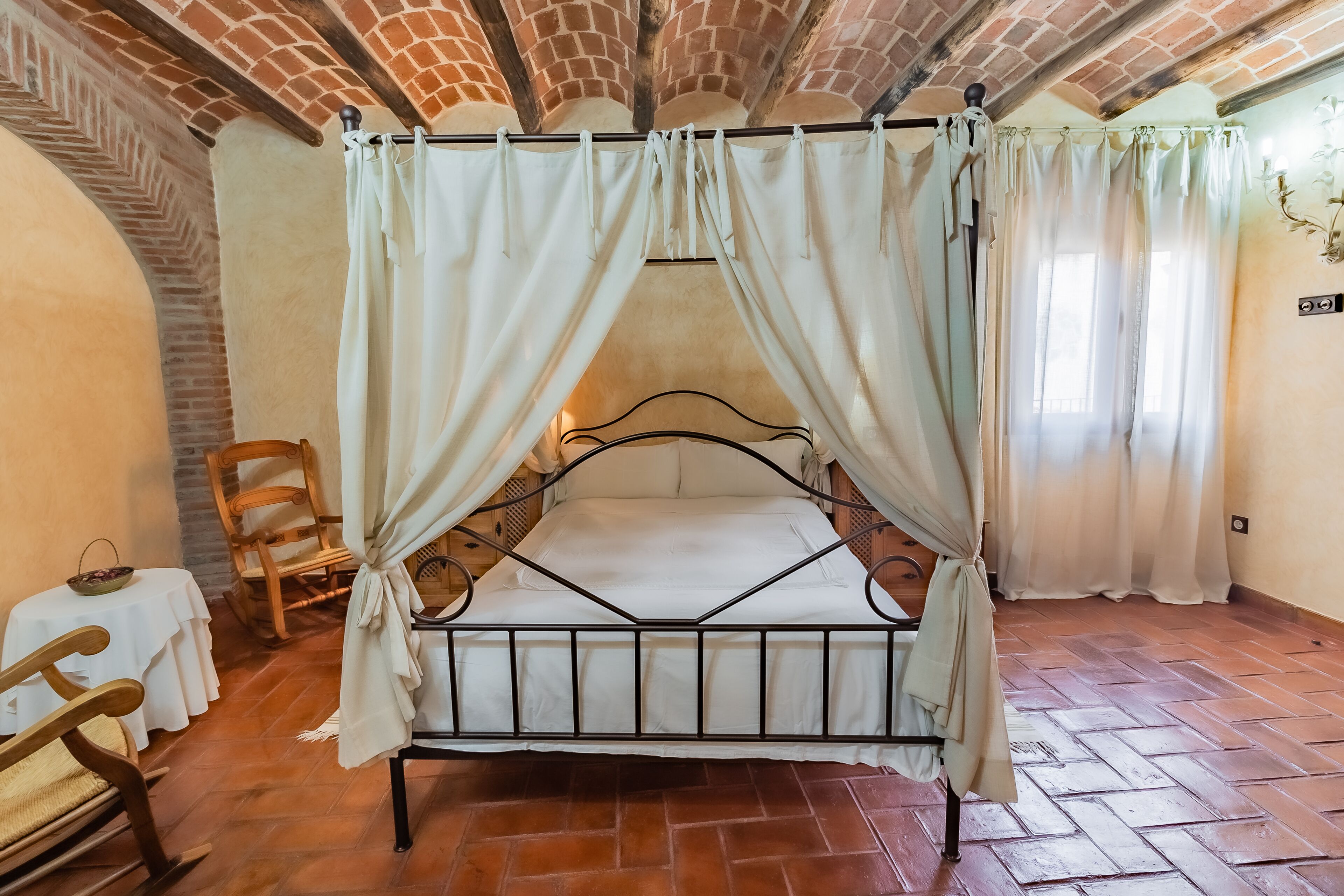 A rustic bedroom with a metal bed, white curtains, a wooden chair, and a small table.