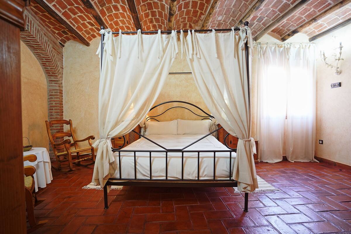 A four-poster canopy bed with a white canopy and a wooden headboard in a room with brick walls and a tiled floor.