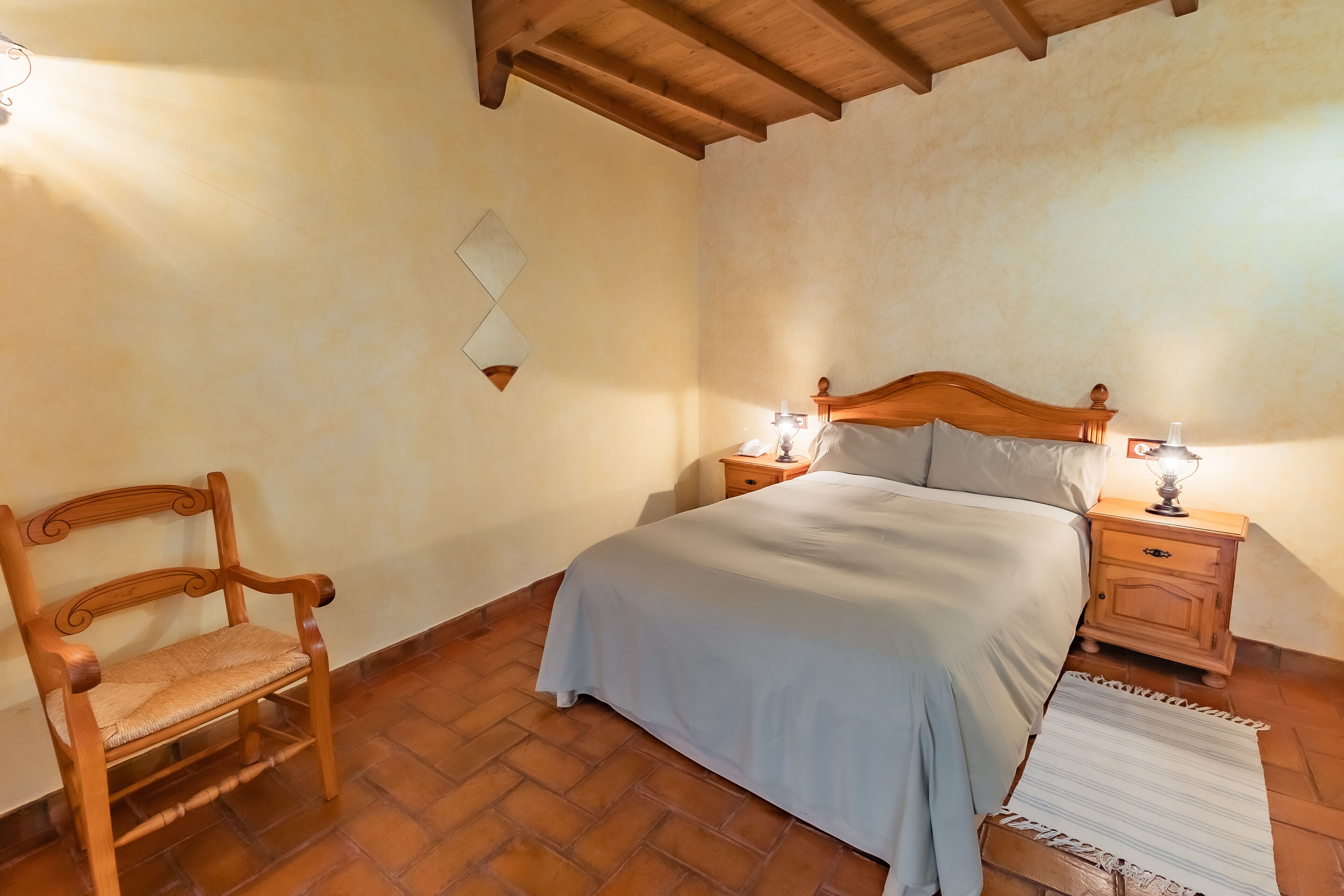 Un dormitorio con cama de madera, dos mesitas de noche con lámparas, una silla de madera y una alfombra estampada sobre piso de baldosas.