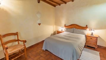 Un dormitorio con cama de madera, dos mesitas de noche con lámparas, una silla de madera y una alfombra estampada sobre piso de baldosas.