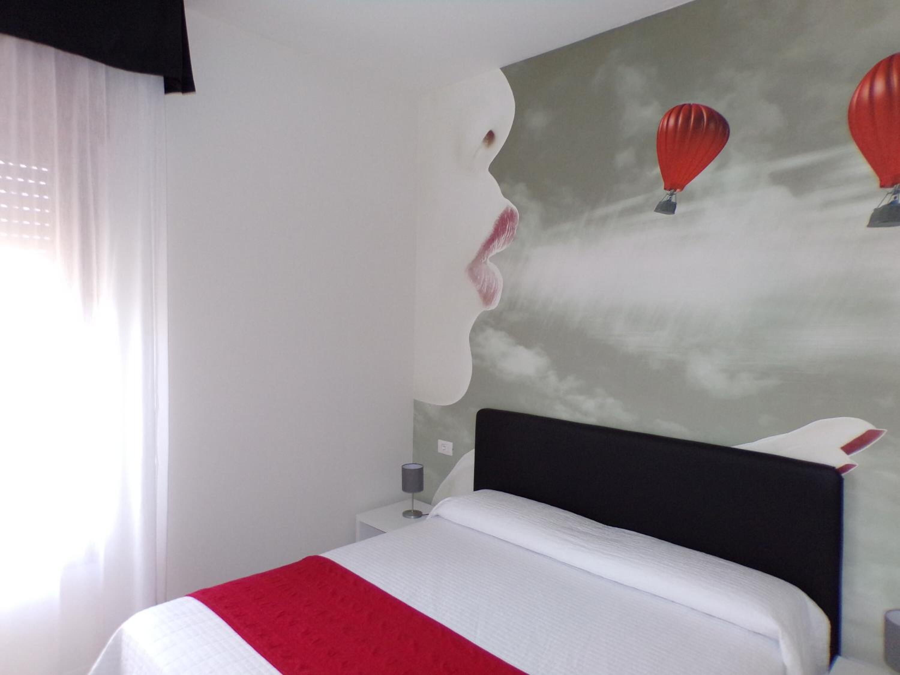 Un dormitorio con cama, mesita de noche, lámpara y una pared con una obra de arte que muestra el perfil de una mujer y globos aerostáticos.