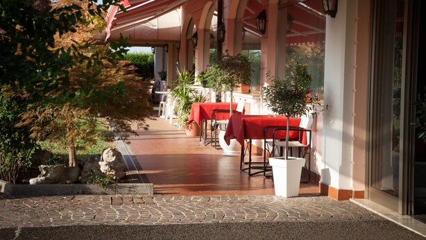 Un patio al aire libre con mesas y sillas, cubierto por un toldo rojo, rodeado de plantas en macetas y un camino de piedra.