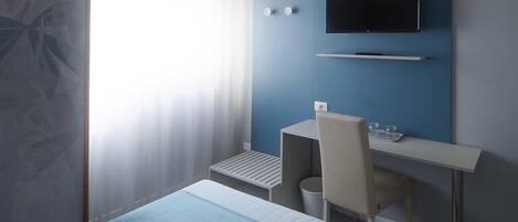 A hotel room with a bed, a desk, a chair, a TV mounted on the wall, and a window with curtains.