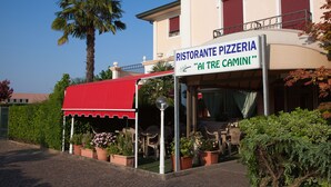 Pizzería con mesas al aire libre, toldo rojo y plantas en macetas.