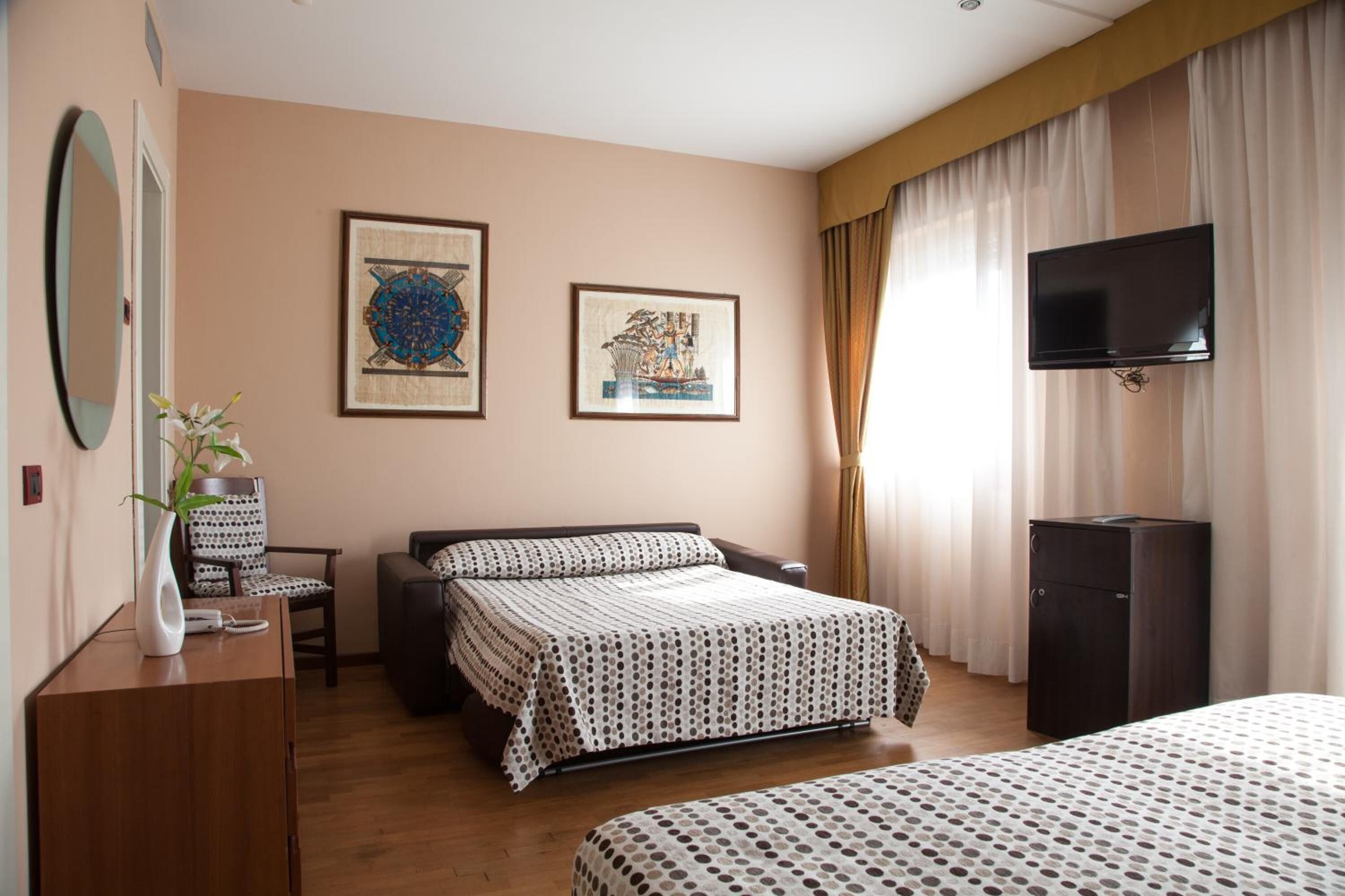 A hotel room with a bed, a desk with a chair, a television, and two framed artworks on the wall.