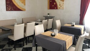 Un comedor con mesas y sillas, dos cuadros en las paredes y una planta en maceta sobre una mesita.