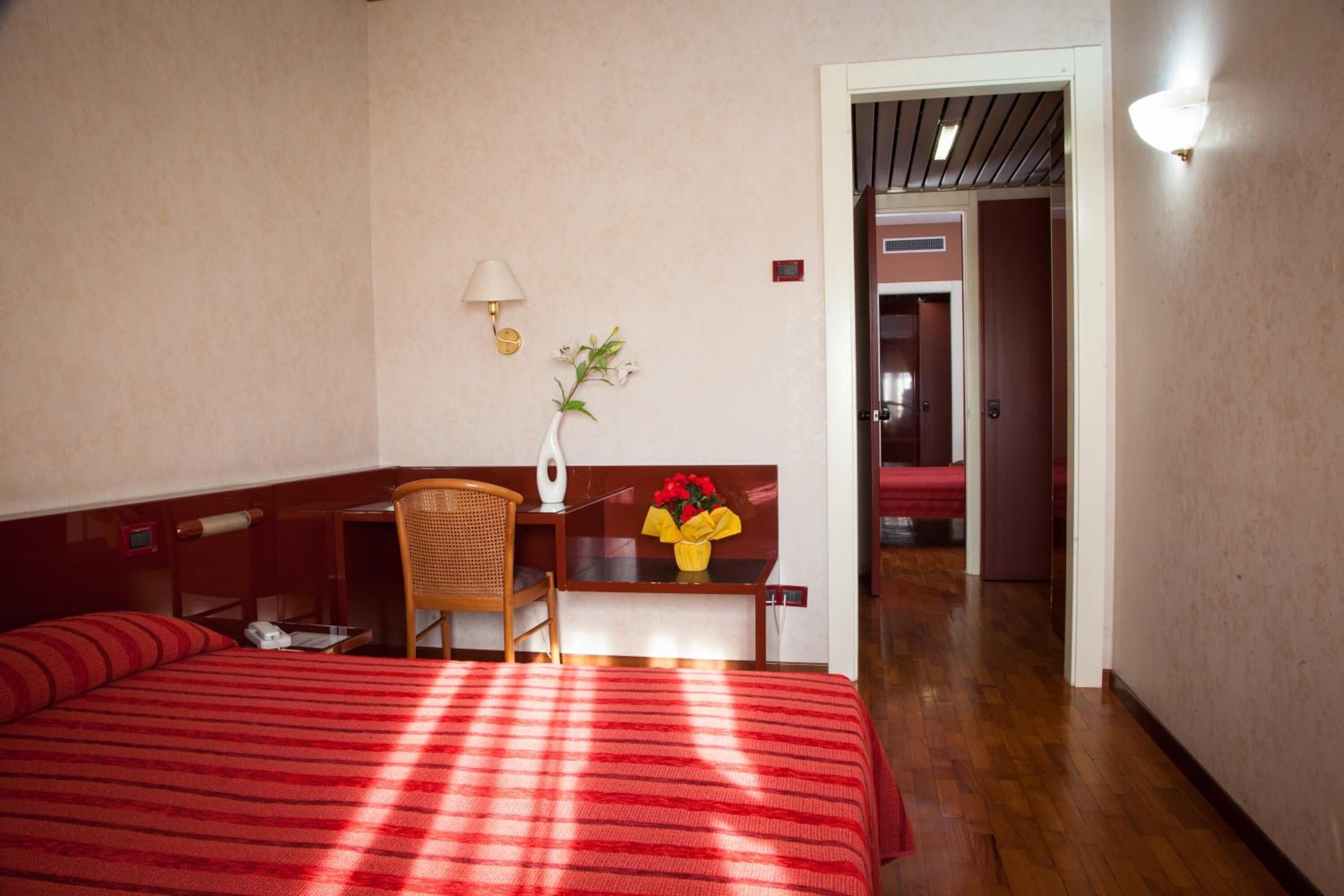 Una camera d'albergo con un letto, una scrivania, una sedia, una lampada e un vaso con fiori.