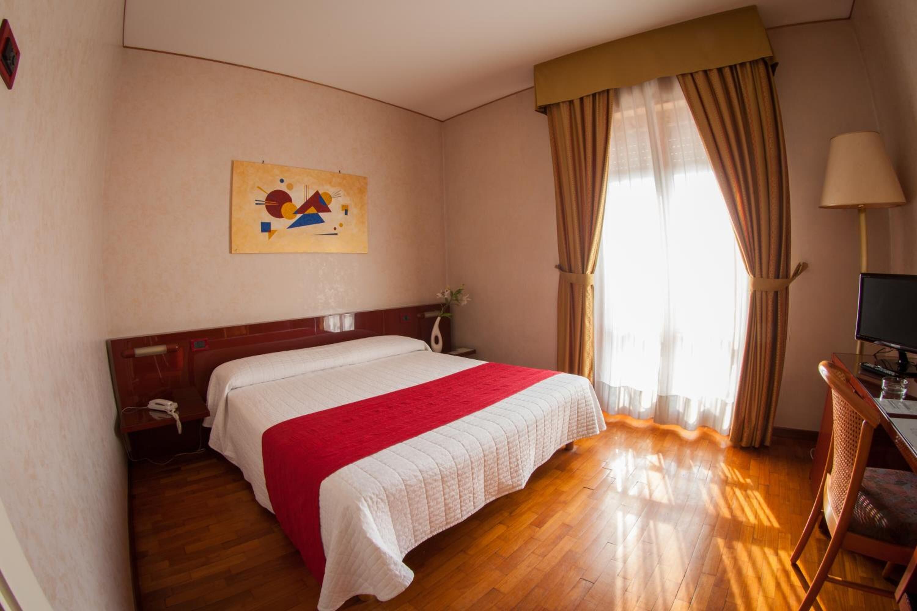 Camera d'albergo con un letto, una scrivania, una sedia, una lampada, un quadro e una finestra con tende.