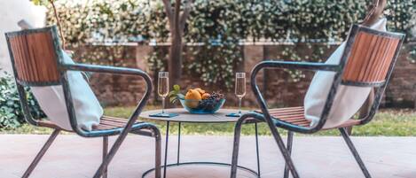 A patio with two metal chairs and a round table, a bowl of fruit, and two glasses of a clear liquid.