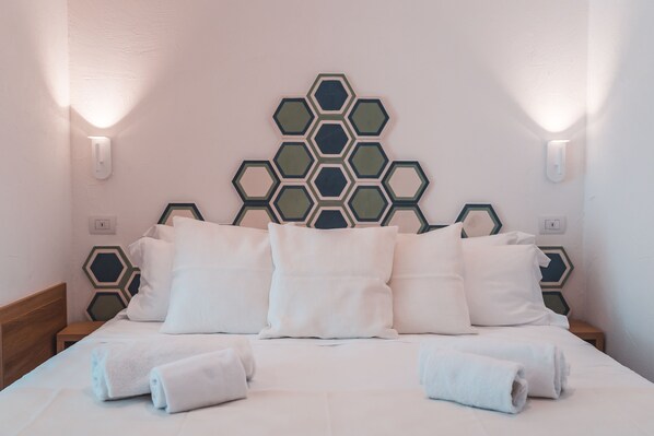 Una cama bien tendida con sábanas y toallas blancas, un adorno geométrico en la pared y dos lámparas fijadas a la pared.