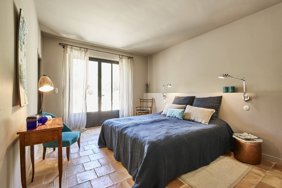 Een slaapkamer met een bed, een bureau, een stoel, een lamp en een raam met gordijnen.