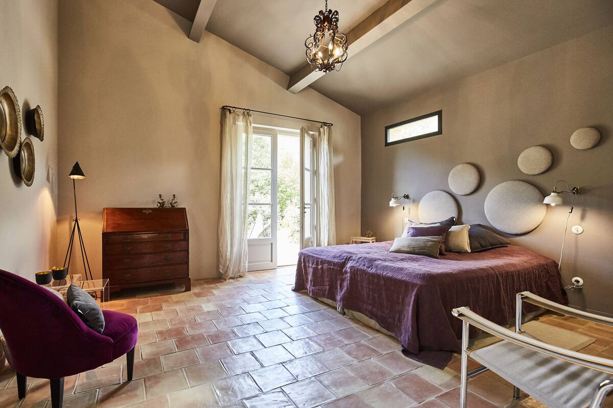 Een slaapkamer met een bed, een dressoir, een stoel, een lamp en een raam met gordijnen.