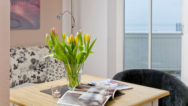 Una habitación con una mesa de madera, un jarrón de vidrio con flores, una vela encendida, una revista y un sofá con tapicería floral.
