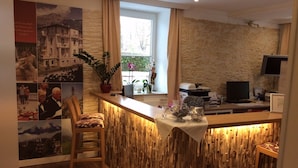 A hotel reception area with a wooden reception desk, a chair, a potted plant, a window with curtains, a stone wall, and a flat-screen TV. - Hotel Almrausch (Bad Reichenhall)