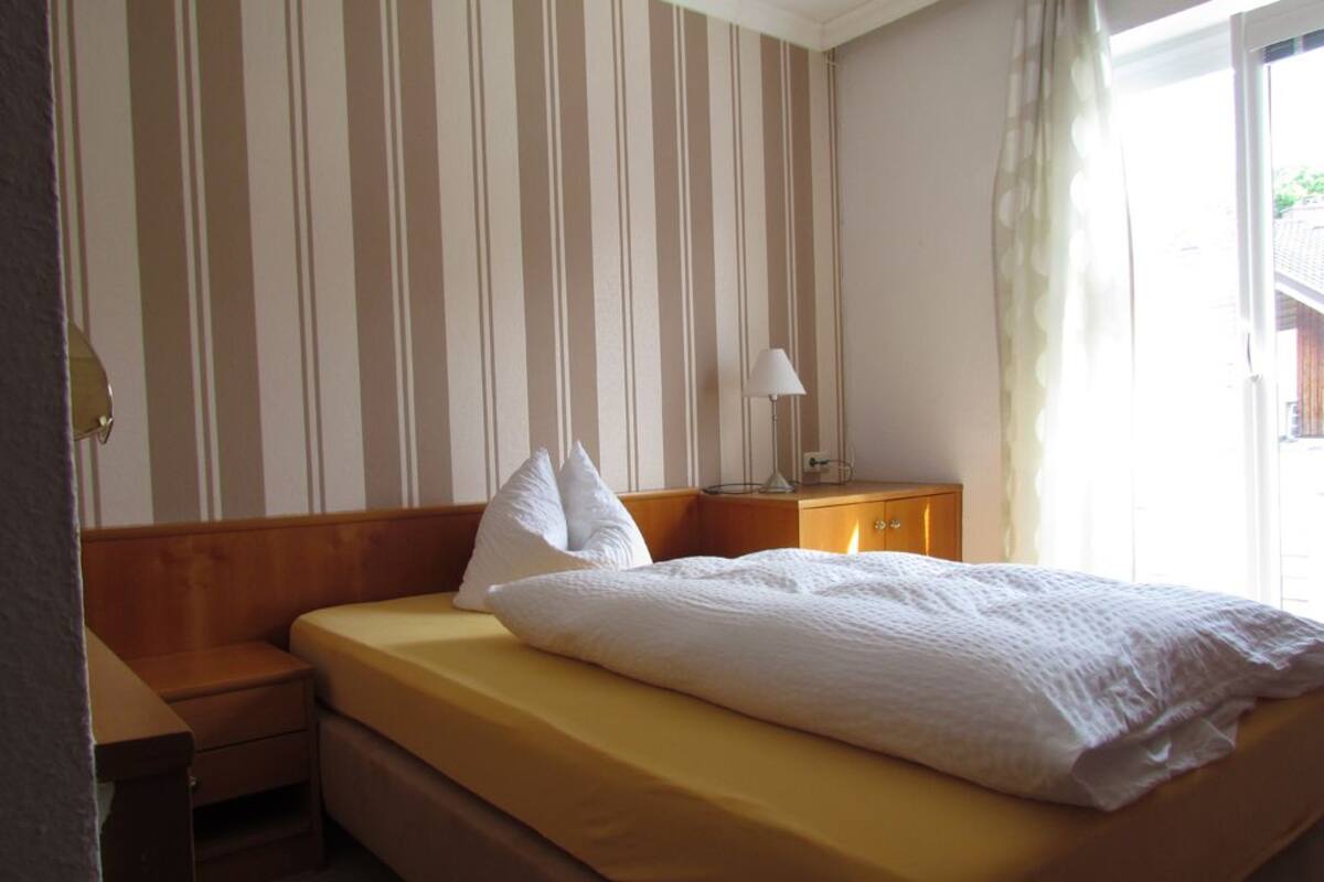 Ein Einzelbett mit Holz-Kopfstück, ein Nachttisch, eine Lampe und ein Fenster mit Vorhängen.