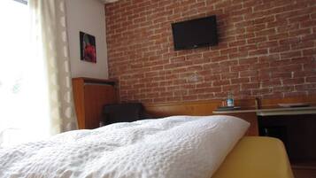 A hotel room with a brick wall, a flat-screen TV mounted above a wooden desk, a bed with a white duvet, and a window with curtains.