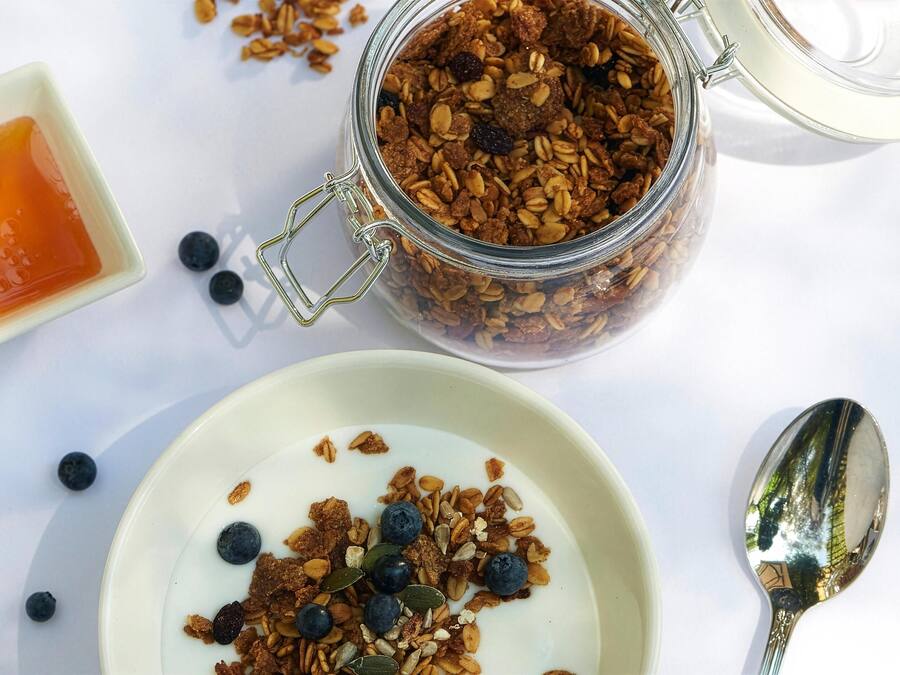 A glass jar filled with granola and blueberries, a small bowl of yogurt, and a spoon on a white surface.