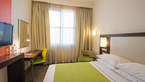 A hotel room with a bed, a desk with a chair, a television, and a window with curtains. - StayEasy Lusaka (Lusaka)