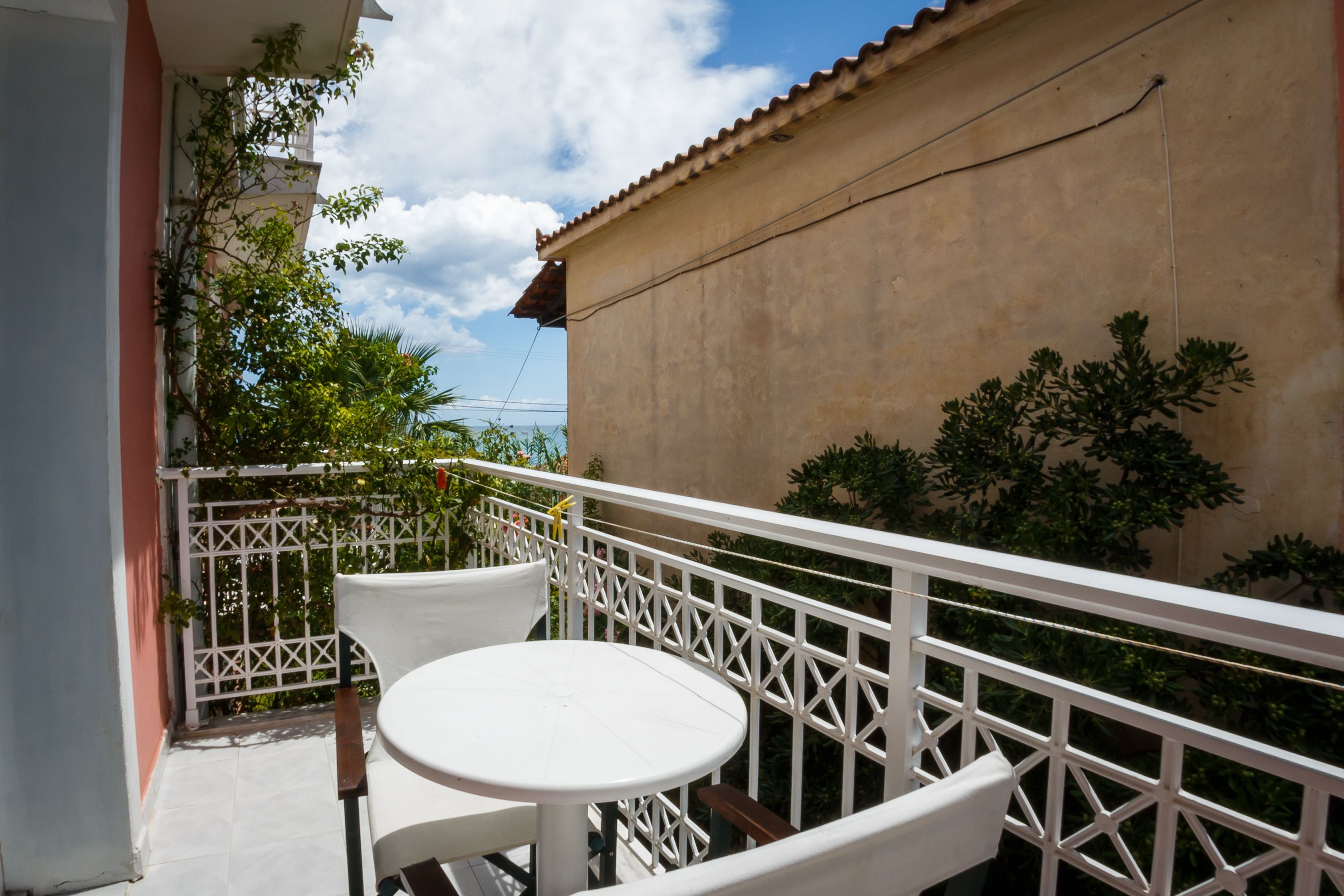 Un balcon avec une table blanche et deux chaises, une balustrade blanche, et un bâtiment avec un toit en tuiles en arrière-plan.