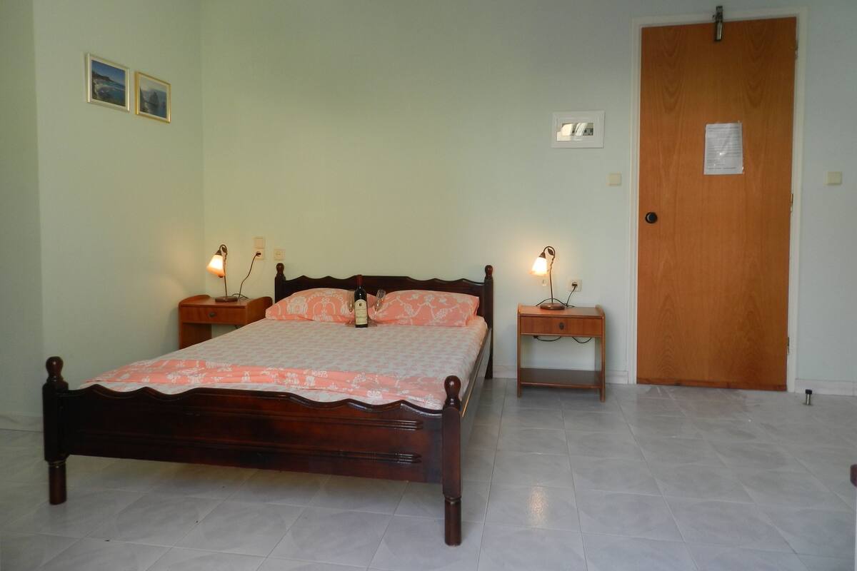 A bedroom with a wooden bed, two bedside tables with lamps, a door, and a light switch on the wall.