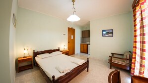 A bedroom with a wooden bed, bedside tables, a nightstand, a chair, a small table, a kitchen area, and a bathroom visible through an open door. - Terra Azzura (Zakynthos)