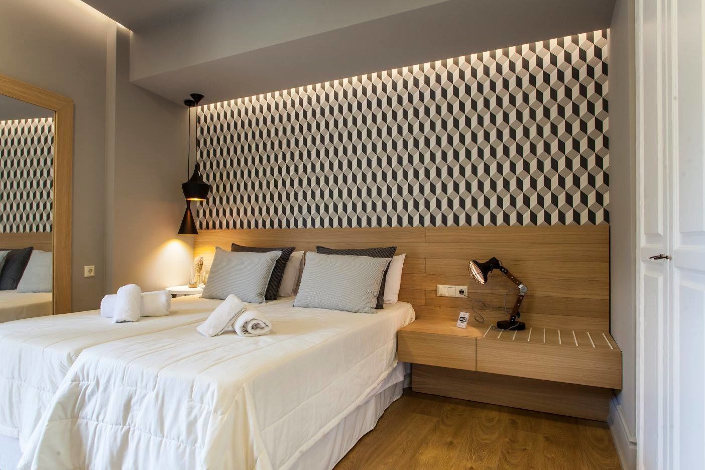 A hotel room with a large bed, a wooden headboard, a bedside table with a lamp, and a wall with a geometric pattern.