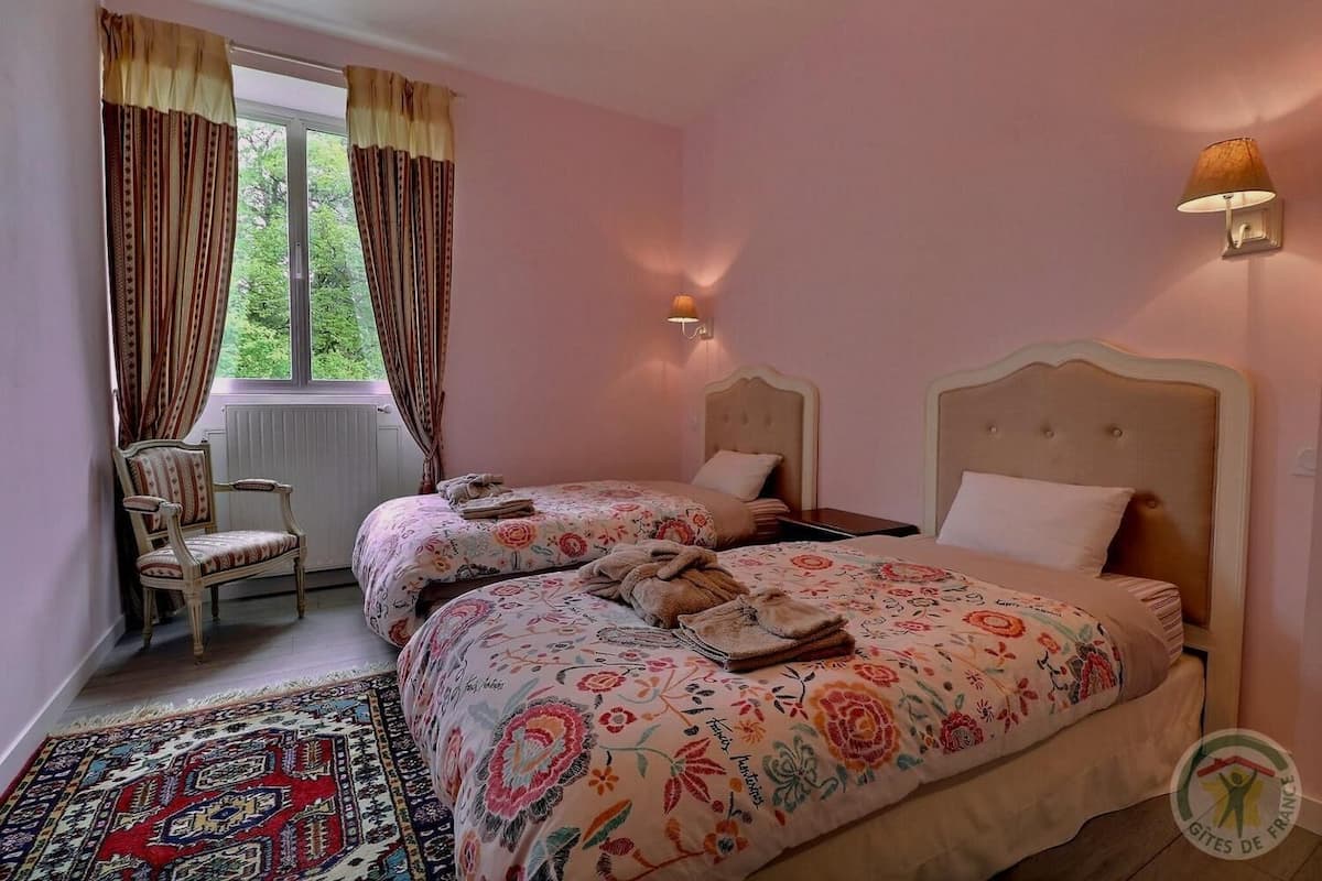 A bedroom with two beds, a chair, a window with curtains, a wall-mounted lamp, and a patterned rug.