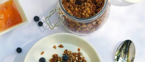 Un frasco de vidrio lleno de granola y arándanos, un tazón pequeño de yogur y una cuchara sobre una superficie blanca.