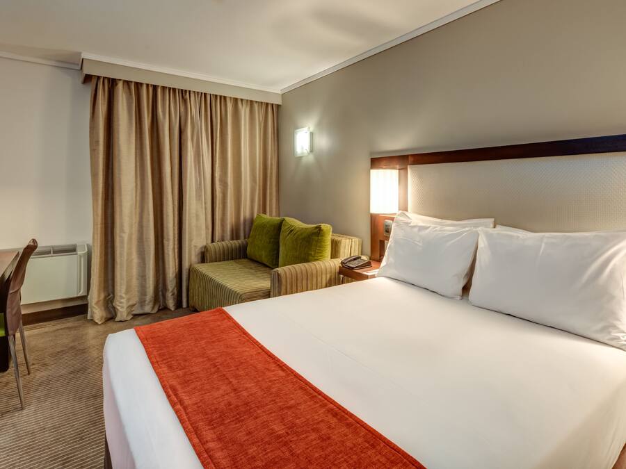 Una habitación de hotel con cama, escritorio, silla, lámpara y ventana con cortinas.