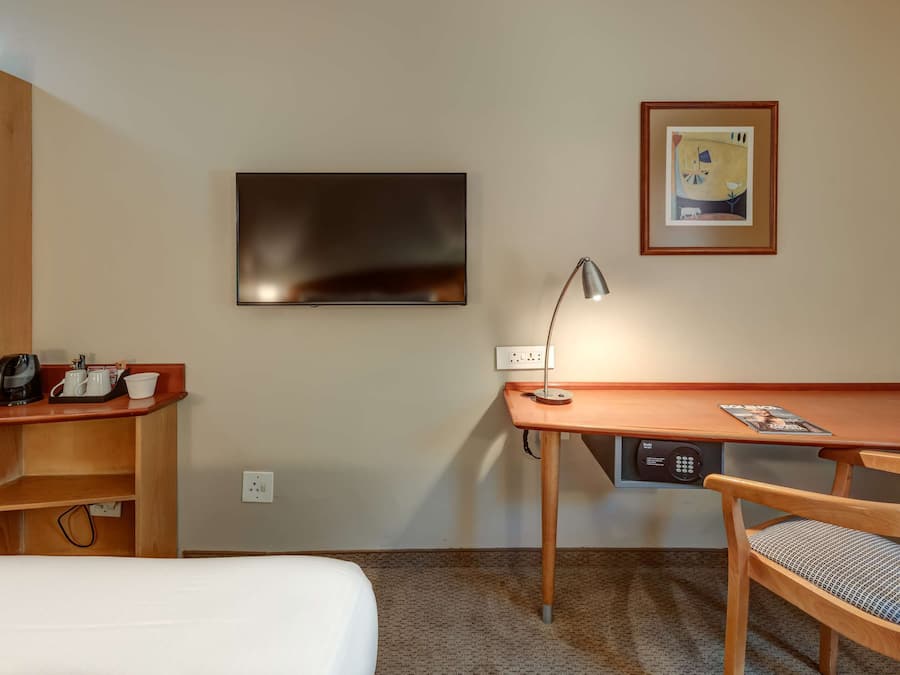 Habitación de hotel con cama, escritorio con lámpara, silla, televisor y un cuadro en la pared.