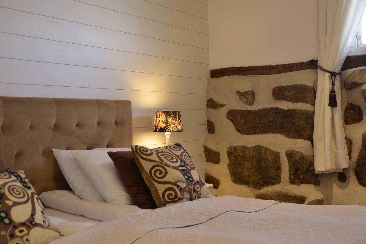 A bedroom with a tufted headboard, patterned pillows, a stone wall, and a lit lamp.