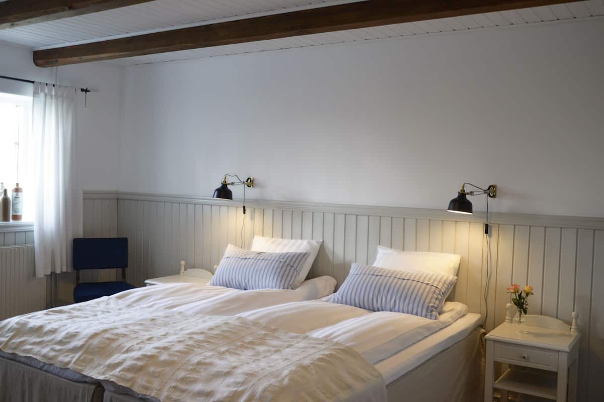 A neatly made bed with white linens, a bedside table with a lamp, and a chair in a room with white walls and wooden beams.