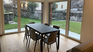 A dining area with a table and chairs, large windows, and a view of a backyard with a stone wall and grass. - Italo-Svizzero (San Giacomo Filippo)
