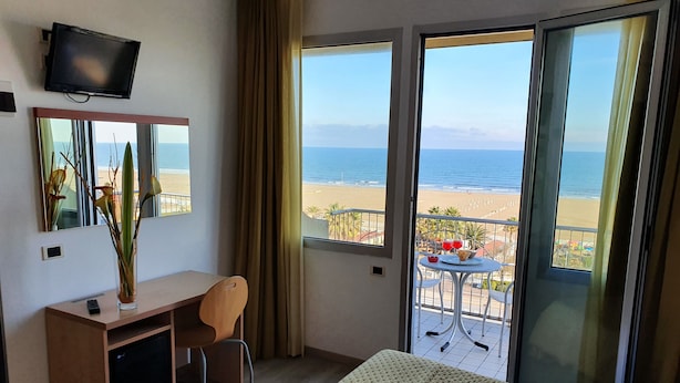 A hotel room with a bed, a desk, a chair, a TV, and a balcony with a view of the beach. - Hotel Tilmar (Rimini)