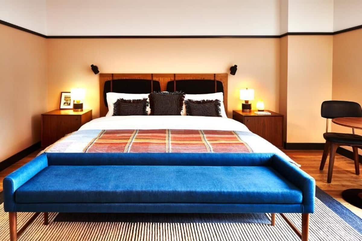 A hotel room with a large bed, two bedside tables with lamps, a blue bench, and a small table with chairs.