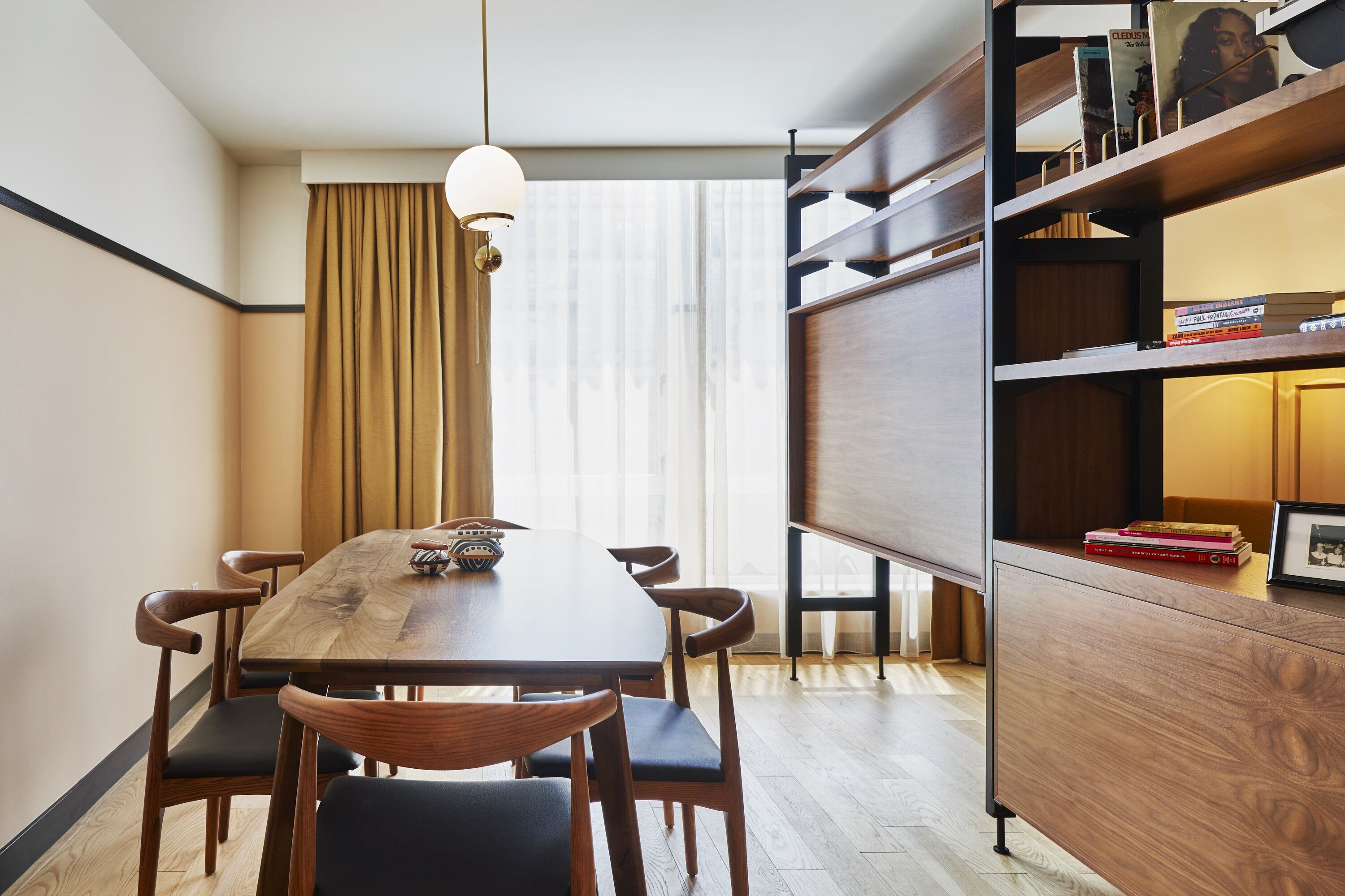 Un comedor moderno con una mesa y sillas de madera, una estantería con libros y objetos decorativos, y un ventanal con cortinas.